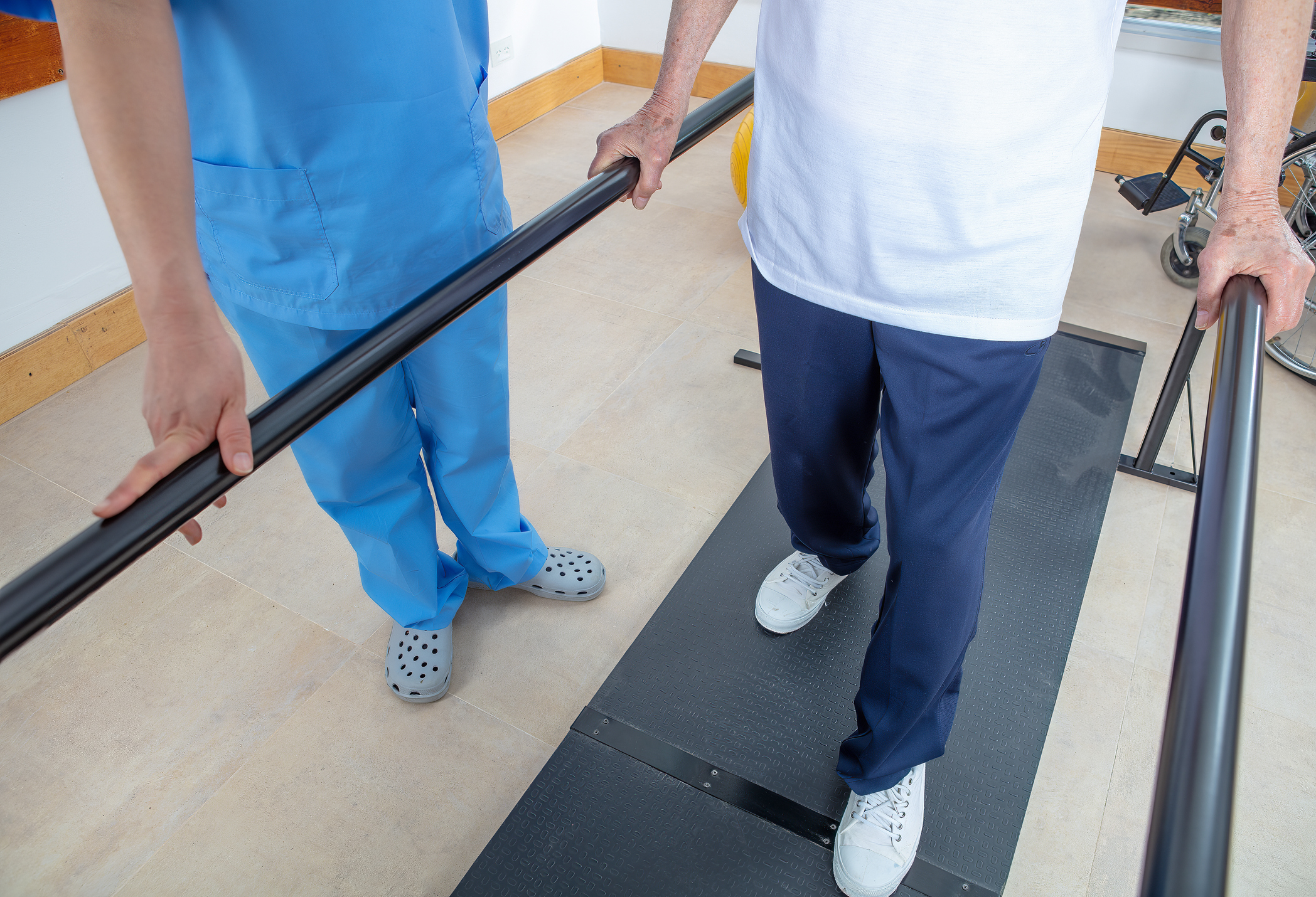person walking with bars in rehabilitation and attended by another person in hospital scrubs